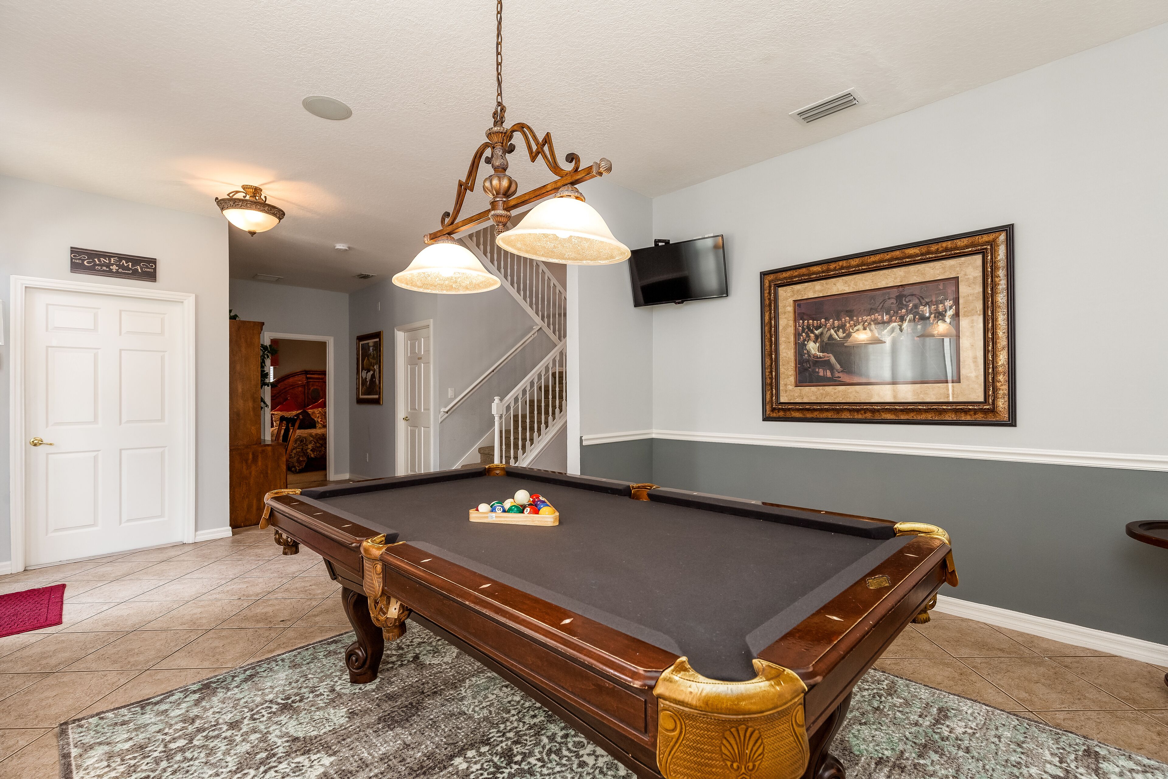 A pool table with a billiard ball rack, a television mounted on the wall, and a staircase in the background.