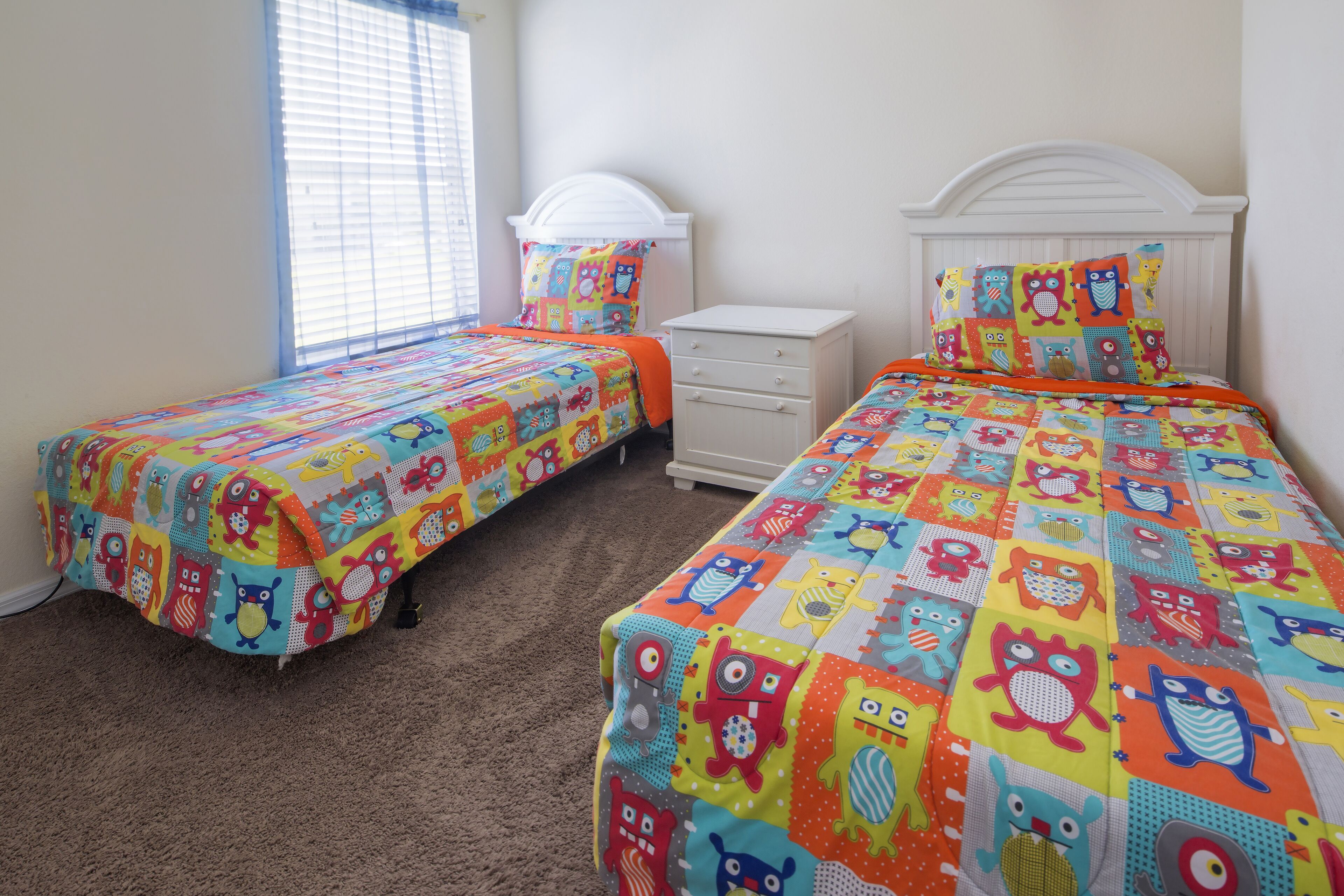 A bedroom with two beds, each with colorful bedding featuring cartoon characters. There is a white dresser and a window with blinds.