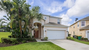 A two-story house with a garage, a well-maintained lawn, and a large tree in the front yard. - Emerald Blossom by VillaDirect (Kissimmee)