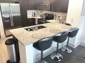 Uma cozinha moderna com bancada de granito, eletrodomésticos de aço inoxidável e banquetas pretas.