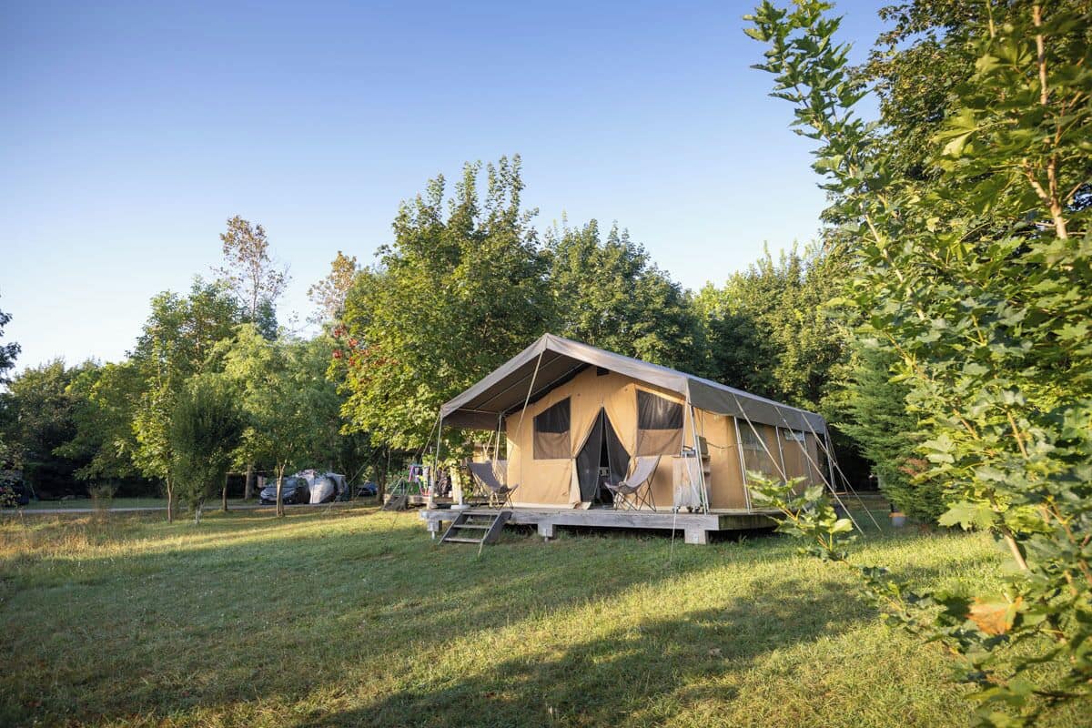 A single-story, A-frame cabin with a flat roof, surrounded by a grassy area and trees. There are two folding chairs outside the cabin.