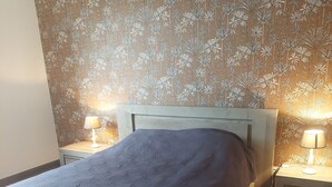 A bed with a headboard, two bedside tables with lamps, and a floral wallpaper pattern. - APARTMENT 4/6 PERS (Oletta)
