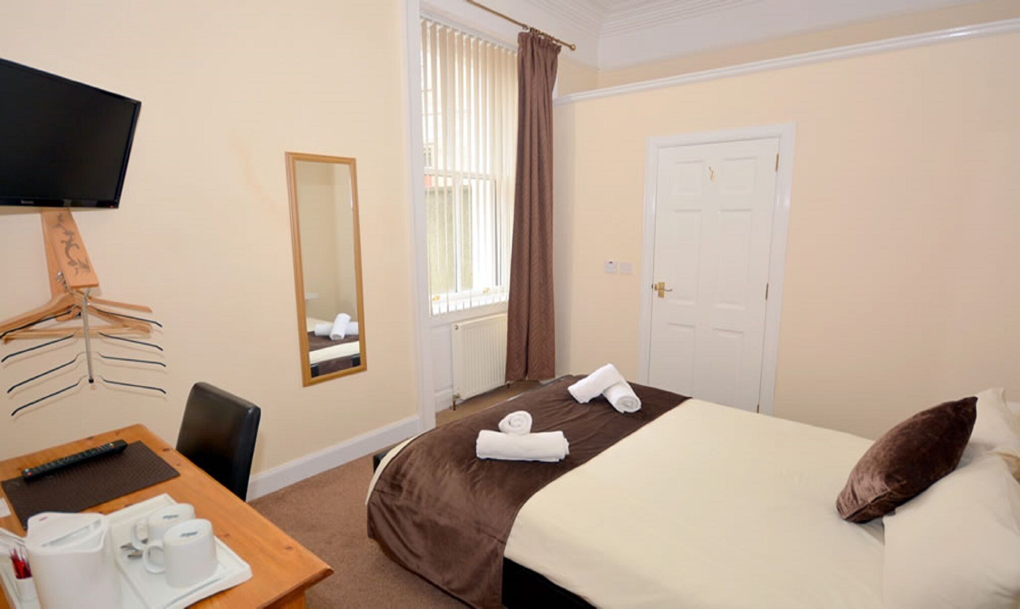 Une chambre d’hôtel comprenant un lit, un bureau avec une chaise, une télévision, un miroir, une porte et une fenêtre avec des rideaux.