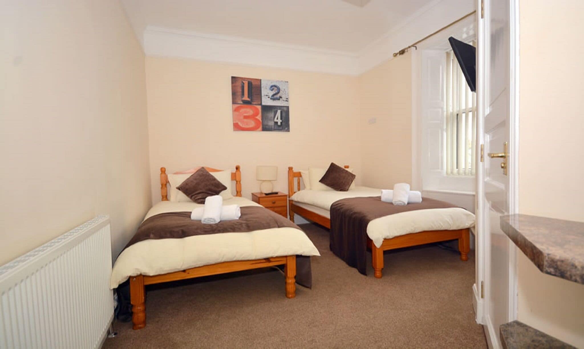 A room with two wooden beds, each with a white and brown bedspread, a nightstand, and a wall clock. There is a window with a curtain and a door with a brass handle.