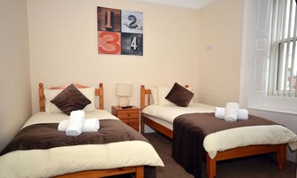 A hotel room with two single beds, each with a brown headboard, white bedding, and a brown throw blanket. There is a nightstand with a lamp and a small table between the beds. A wall-mounted artwork displays the numbers 1, 2, 3, and 4.