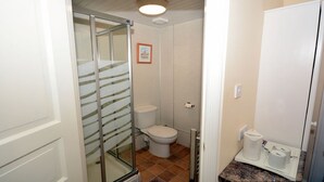 Un baño con cabina de ducha de vidrio, inodoro y lavamanos con cafetera y toallas de papel.