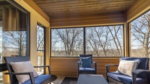 A covered porch with wicker furniture, including two chairs and a small table, and large windows offering a view of bare trees. - The Cozy Lake Stay You've been Craving - Fireplace, Comfy Beds & Quiet! (Galena)