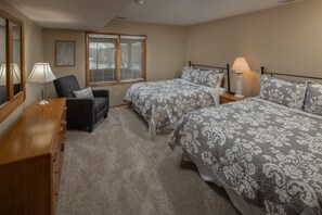 Une chambre avec deux lits, une commode en bois, une chaise, une table de chevet avec une lampe et une fenêtre avec des stores.