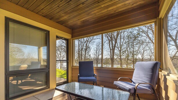 A covered porch with a glass table and two chairs, large windows offering a view of trees and a lawn. - The Cozy Lake Stay You've been Craving - Fireplace, Comfy Beds & Quiet! (Galena)