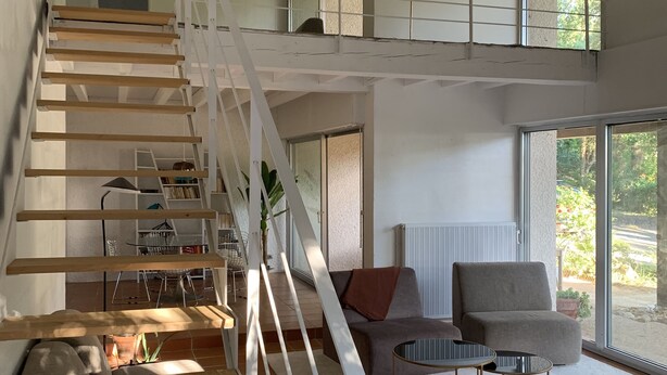 Un vaste espace de vie agrémenté d’un escalier en bois, d’un canapé gris, d’une table basse et offrant une vue sur l’extérieur grâce à de grandes fenêtres.