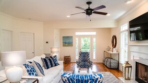 A living room with a white sofa, patterned armchair, and a coffee table. There is a fireplace, a TV, and a ceiling fan. - Beautiful Grand Pavilion -  steps to the beach with Grand Pavilion Pool Access (Isle of Palms)