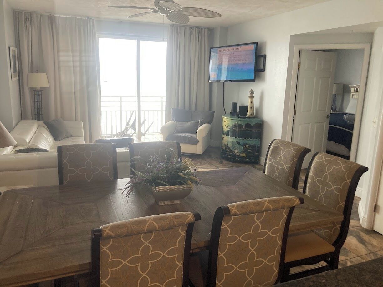 A dining area with a wooden table and patterned chairs, a television on the wall, and a view of the outdoors.