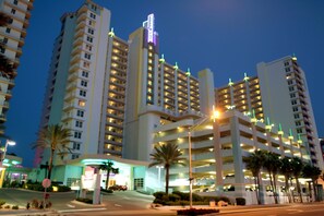 Um hotel com vários andares, um letreiro iluminado em destaque, várias estruturas de estacionamento e palmeiras em primeiro plano.