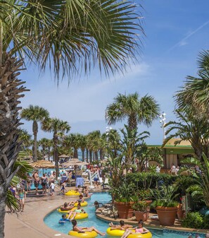 A pool area with multiple lanes for water activities, surrounded by palm trees and shaded by large umbrellas.