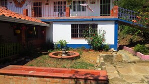 Una casa de dos pisos con fachada azul y blanca, un pequeño jardín con una fuente y un balcón con barandilla.