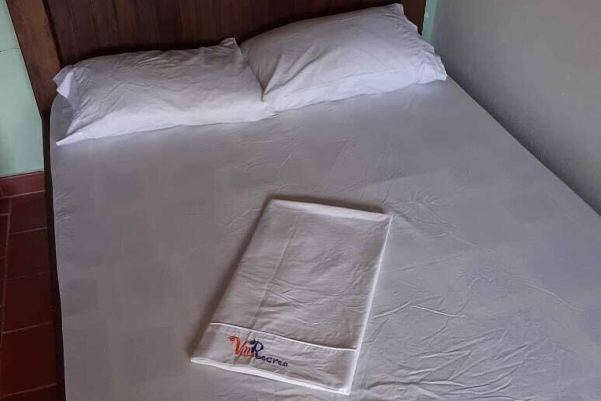 Un lit bien fait, avec un couvre-lit blanc et des oreillers, une tête de lit en bois et un petit objet rectangulaire posé sur le lit.