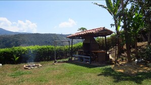 Un espace extérieur rustique aménagé avec un coin salon, un barbecue en briques et une balançoire, le tout entouré d’une végétation luxuriante et dominé par un paysage montagneux.