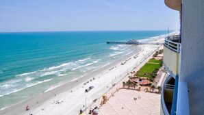 A beach view from a balcony with a pier in the distance, a sandy beach, and a paved area with a circular structure.