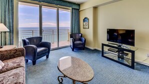 A hotel room with a large sliding glass door leading to a balcony, a flat-screen TV, two leather armchairs, a patterned sofa, and a small table.