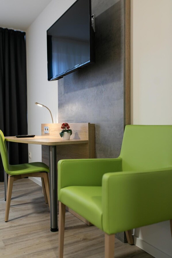 A modern room with a flat-screen TV mounted on the wall, a wooden table with a lamp, a small plant, and two green chairs. - Gasthaus zur Blume (Efringen-Kirchen)