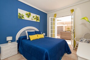 A bedroom with a blue bedspread, white headboard, and yellow pillows. There is a white nightstand with a lamp and a white dresser. A painting of flowers hangs on the wall, and a plant is on the windowsill.