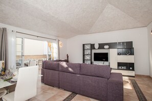 A modern living room with a grey sofa, a dining area with a table set for two, and a TV mounted on the wall.