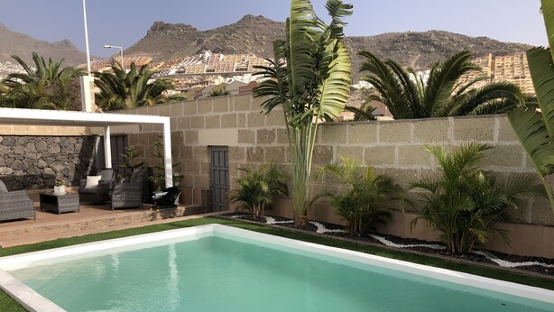Una piscina con un muro de piedra y plantas tropicales, un área de descanso cubierta y vistas a un pueblo montañoso al fondo.