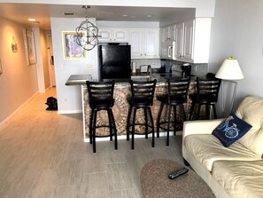 Et moderne køkken med hvide skabe, et sort køleskab og en bar med fire barstole. En beige sofa med en blå pude og en fjernbetjening ligger på et tæppe.