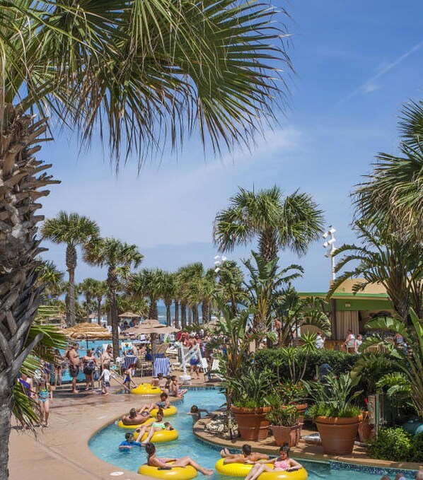 A pool area with multiple lanes for water activities, surrounded by palm trees and shaded by large umbrellas.