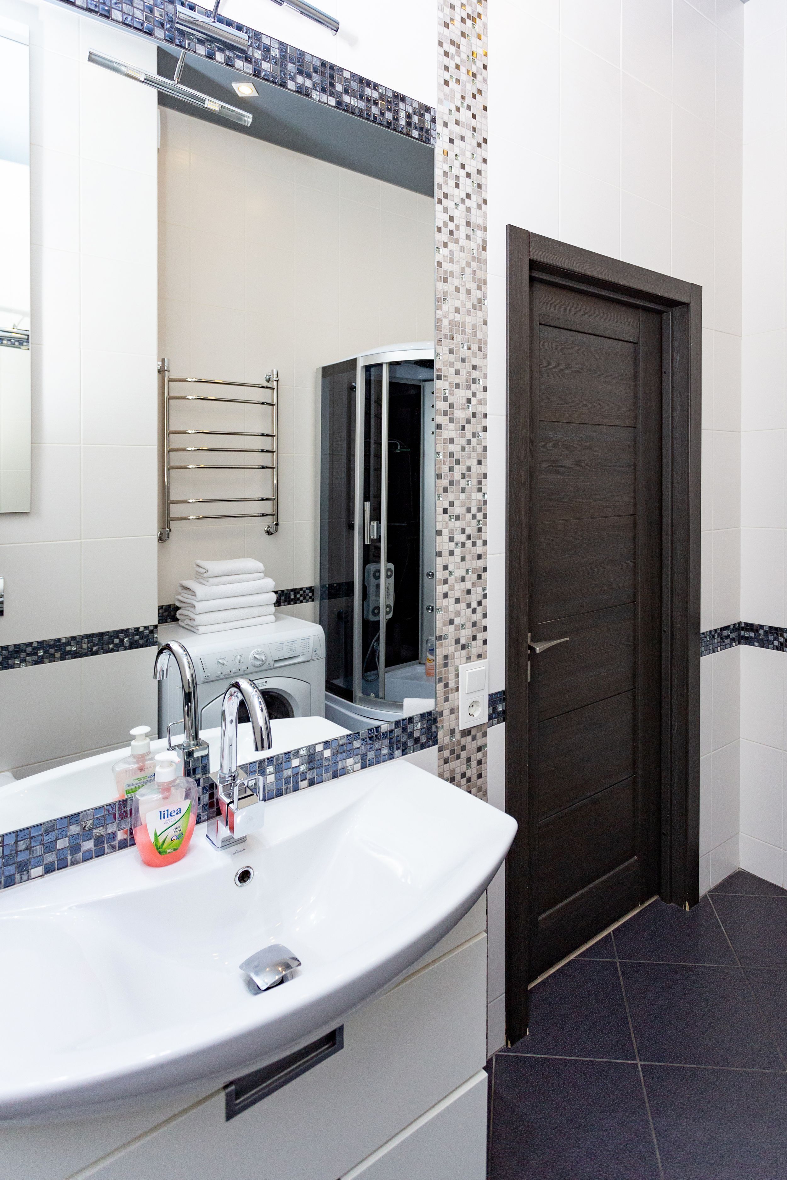 Un baño con un lavamanos blanco, un espejo, un perchero para toallas y una puerta de madera oscura.