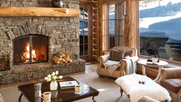 Acogedor interior de cabaña de madera con chimenea de piedra, muebles de madera y ventanas amplias con vistas a la montaña.