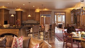 Una cocina amplia con armarios de madera, una isla central e iluminación colgante. Cuenta con una zona de comedor con mesa y sillas, y un sofá de cuero con cojines estampados.