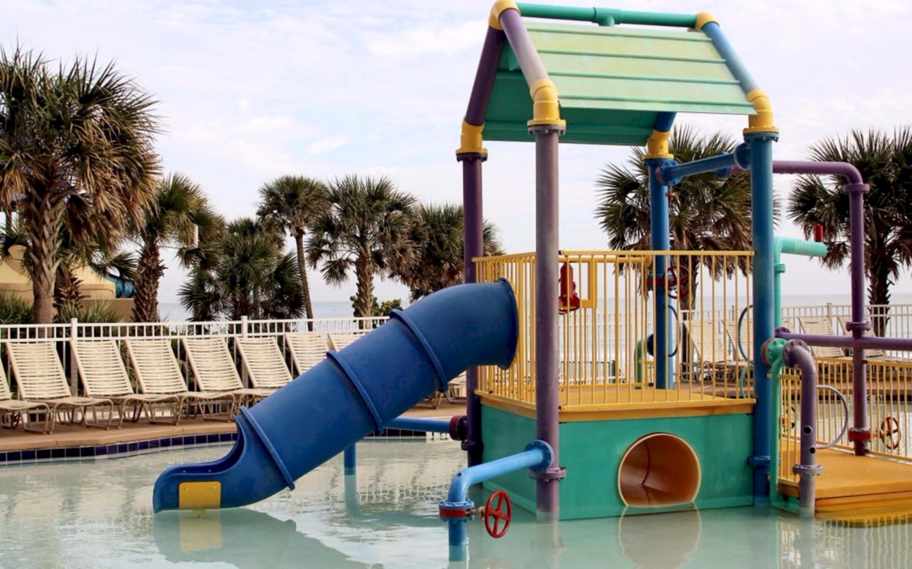 A water play area with a slide and a small pool, surrounded by palm trees and lounge chairs.