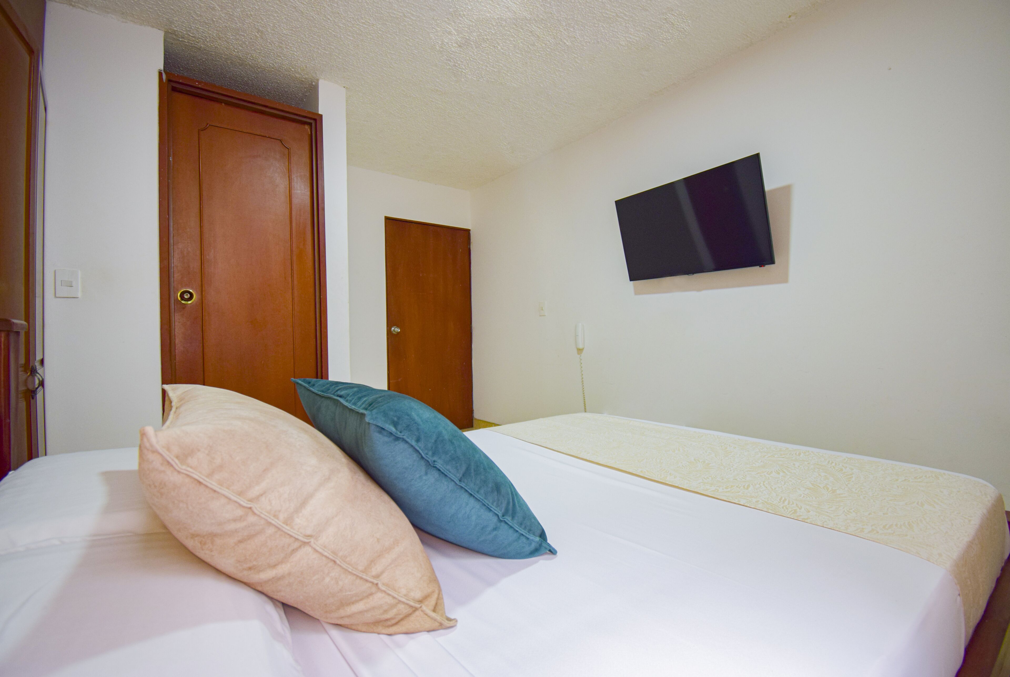 Een slaapkamer met een bed, twee kussens, een aan de muur bevestigde televisie en twee deuren.