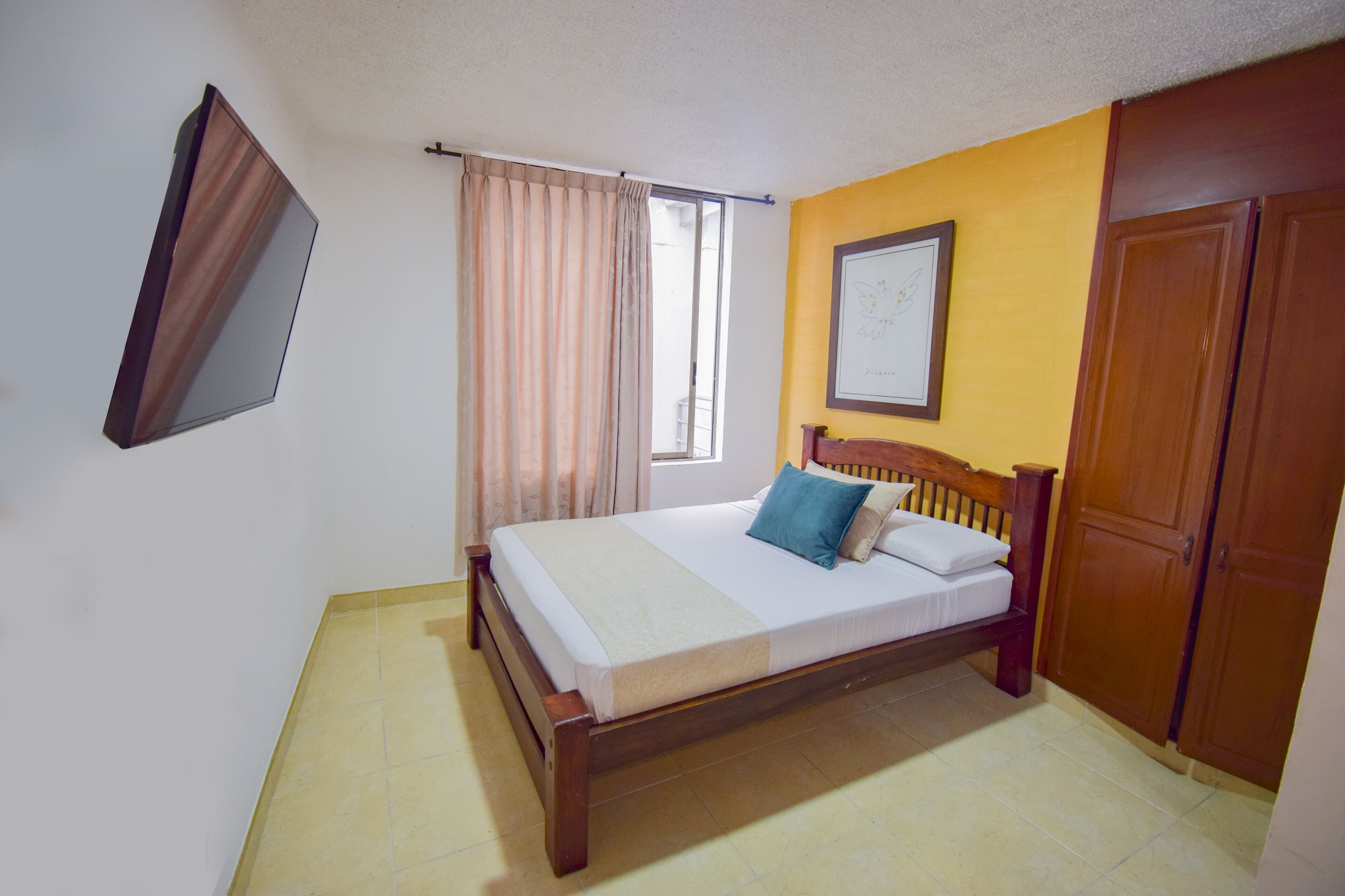 Une chambre à coucher avec un lit, une fenêtre avec des rideaux, une armoire en bois et un téléviseur fixé au mur.