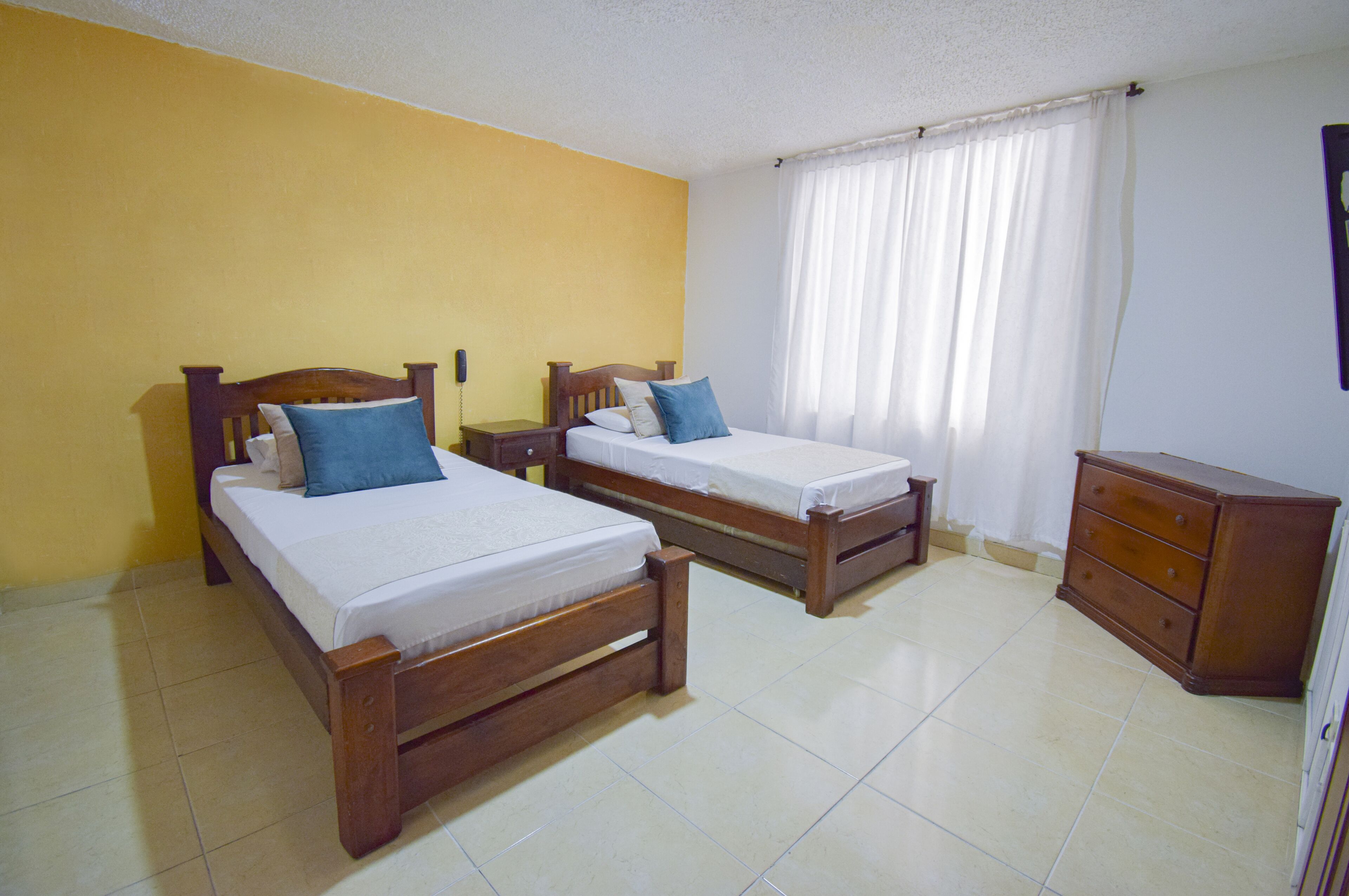 Una habitación con dos camas de madera, una mesita de noche, un aparador y una ventana con cortinas.