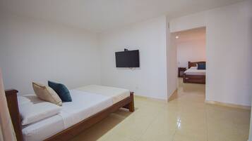 A single bed with a wooden frame and white bedding, a flat-screen TV mounted on the wall, and a tiled floor.