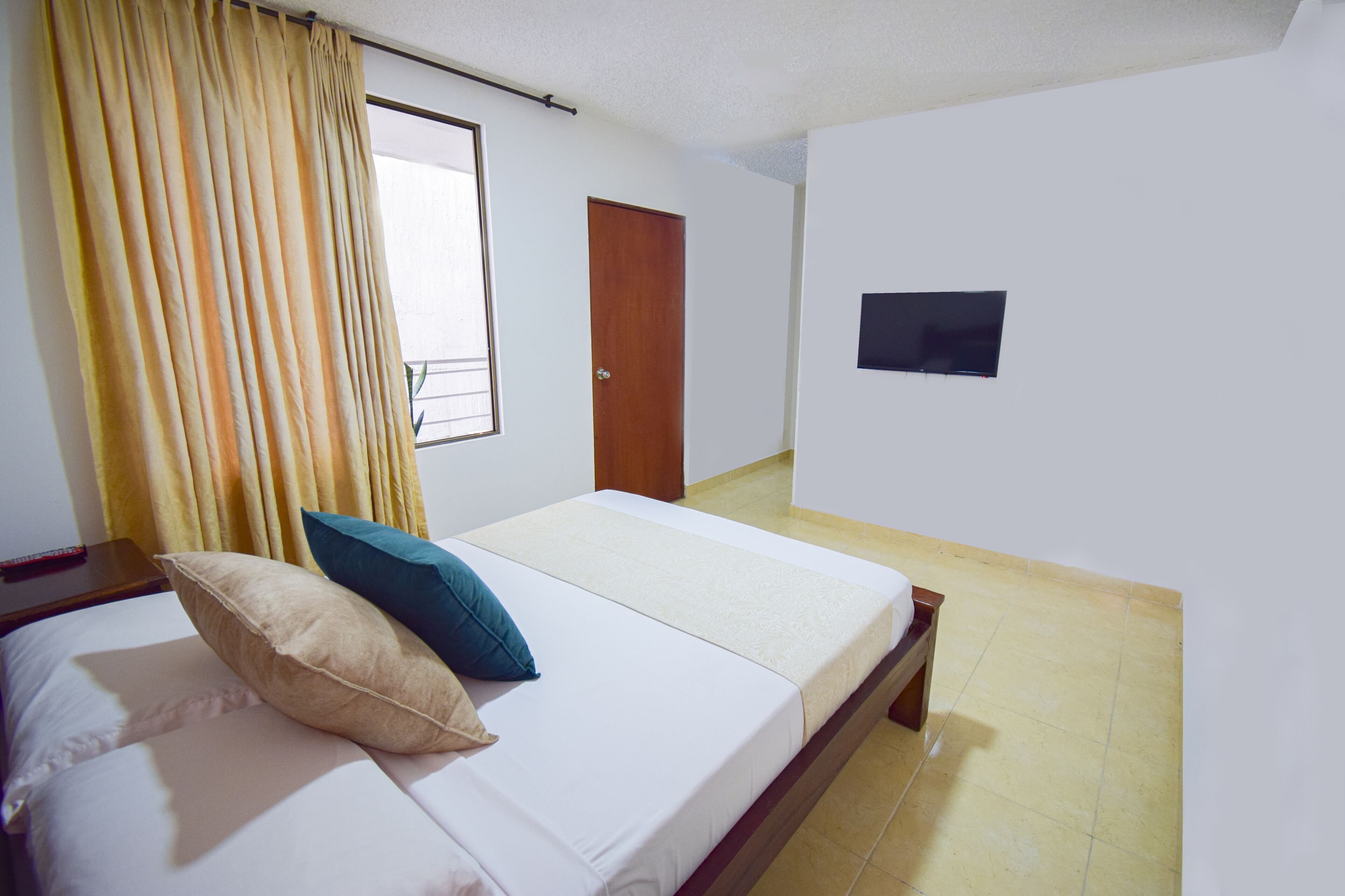 A bedroom with two beds, a window with curtains, a door, and a wall-mounted TV.