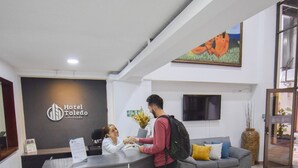 Eine Hotelrezeption mit Rezeptionist, Gast und moderner Einrichtung, darunter ein graues Sofa und ein Flachbildfernseher.