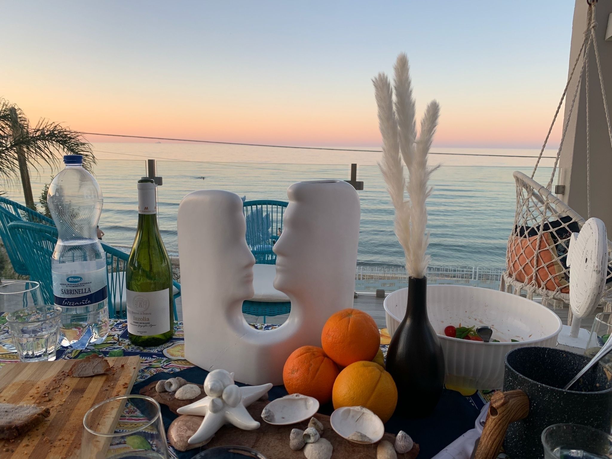 Une table dressée avec une bouteille de vin, des verres, des fruits et des objets décoratifs, le tout placé devant une vue sur la mer.