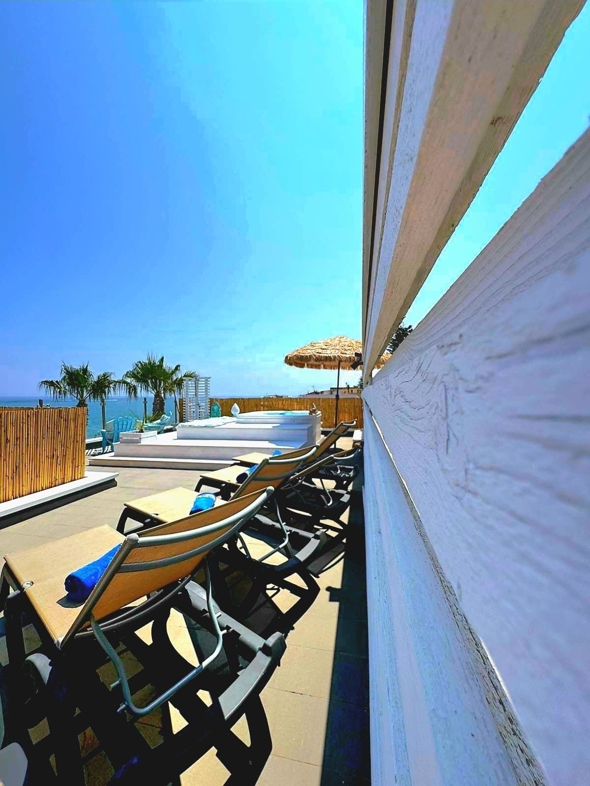Un espace aménagé au bord de la piscine, avec des chaises longues et un jacuzzi, offrant une vue imprenable sur un ciel bleu azur et l’océan.