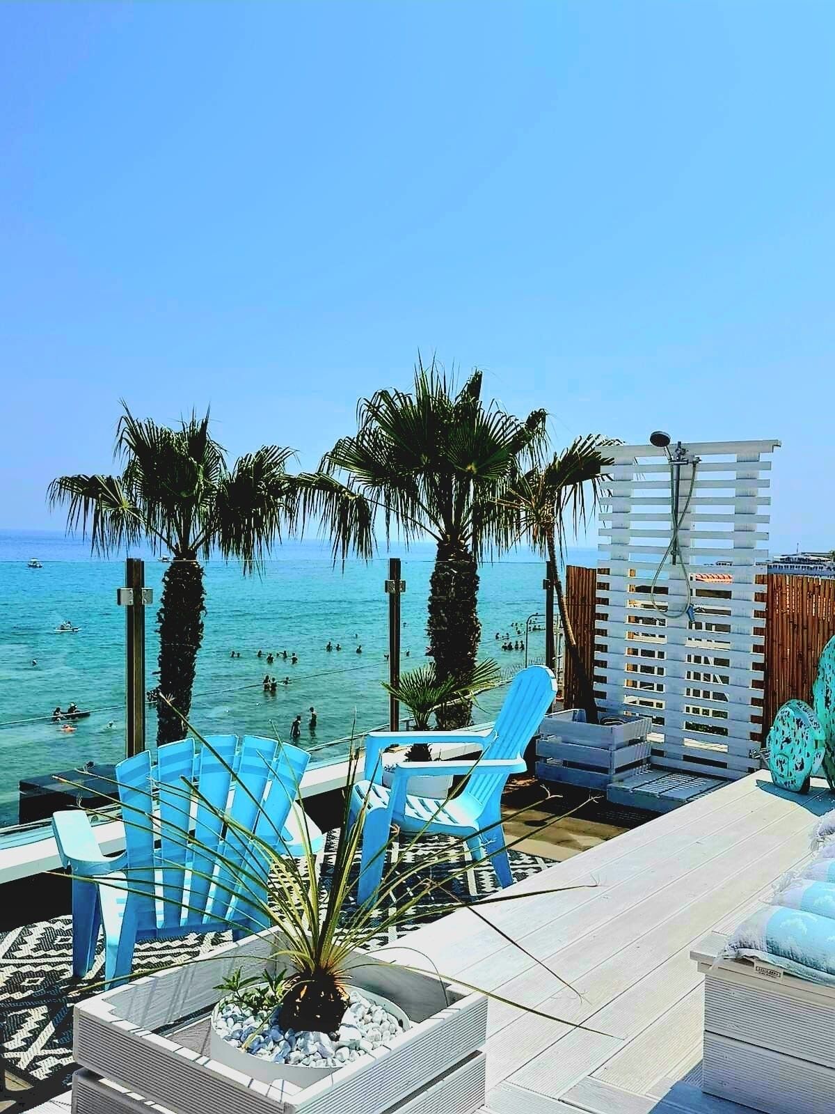 Eine Strandlounge mit blauen Stühlen, Palmen und Blick aufs Meer.