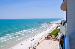 Vista a la playa desde un balcón, con un muelle a lo lejos, arena y una zona pavimentada con una estructura circular.