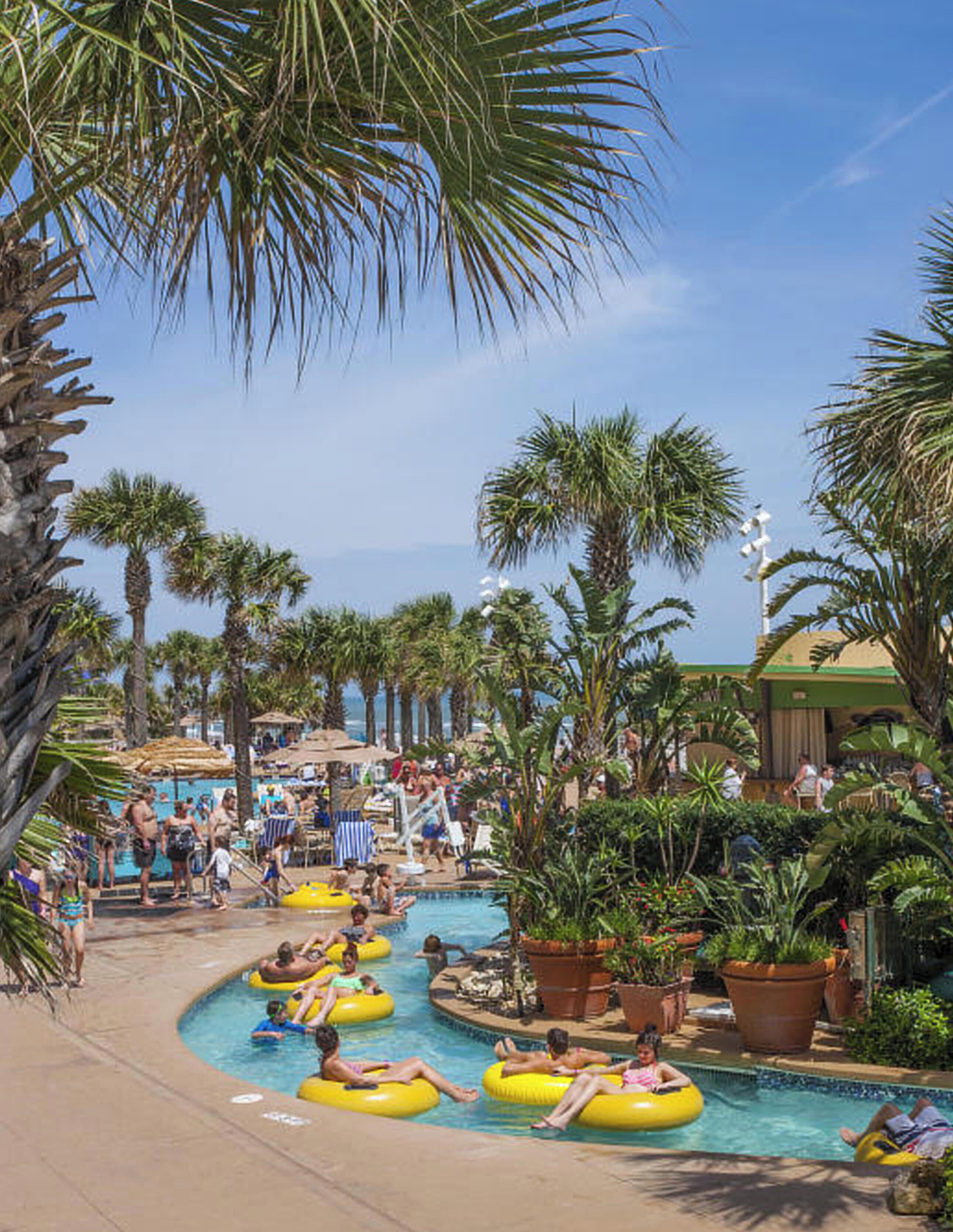 A pool area with multiple lanes for water activities, surrounded by palm trees and shaded by large umbrellas.