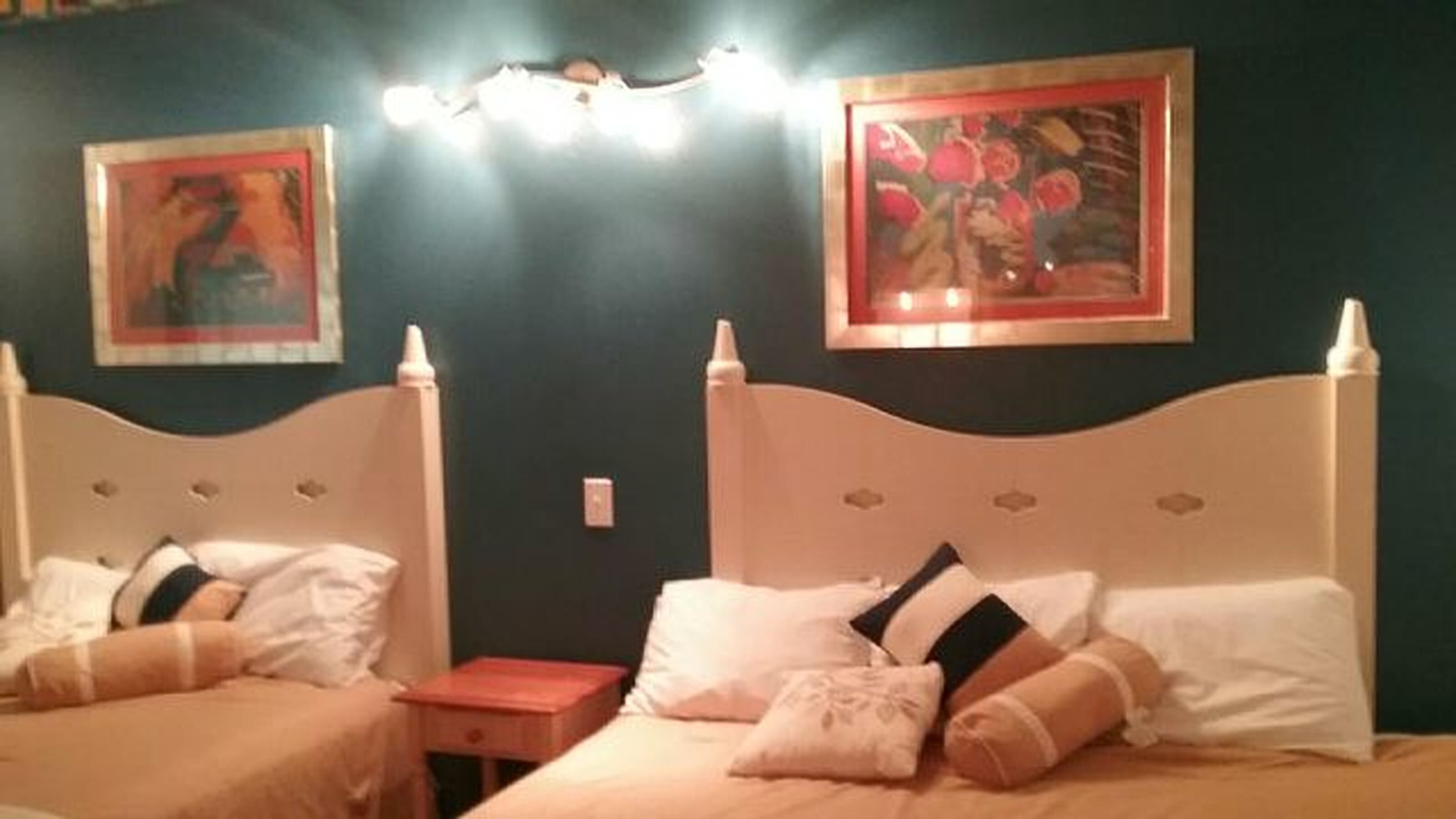 Een kamer met twee bedden, elk met een witte hoofdbord en meerdere kussens. Boven de bedden zijn wandlampen en er hangen twee kunstwerken aan de muur.