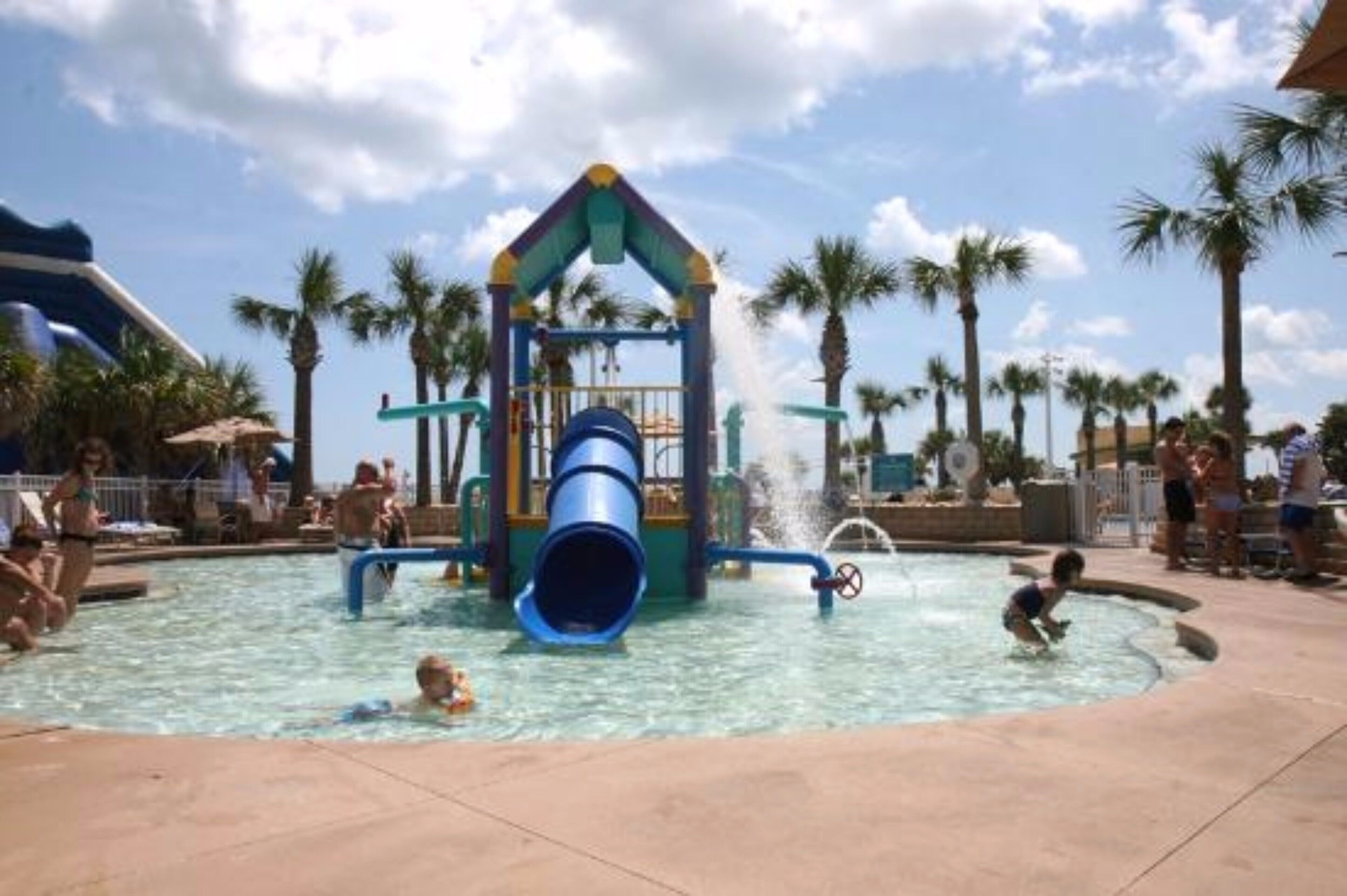 Un espace de jeux aquatiques avec un toboggan et une zone d’éclaboussures, entouré de palmiers et bordé d’une piscine.