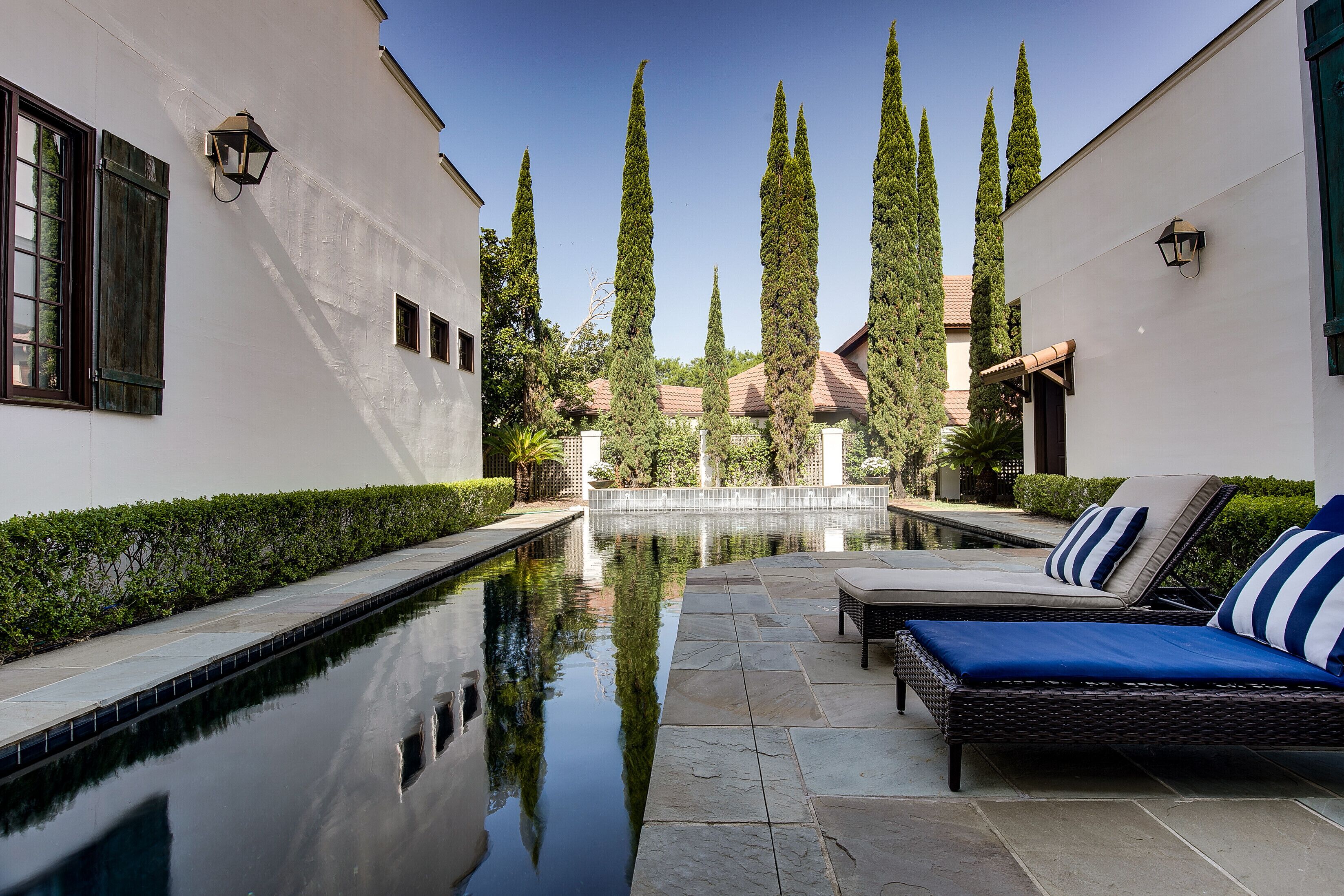 A pool area with a lounge chair and a small table, surrounded by tall cypress trees and a white building with windows.