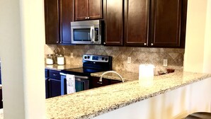 A kitchen with dark wooden cabinets, a stainless steel microwave, a double oven, granite countertops, and two bar stools.