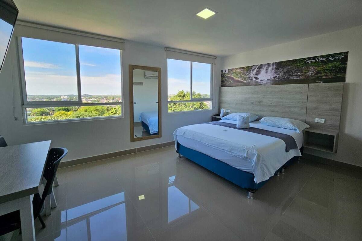 A modern bedroom with a large bed, a desk, and a view of greenery through the windows.