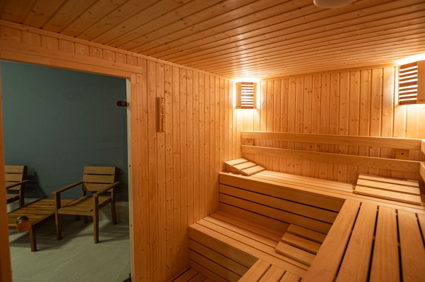 Uma sauna com bancos e paredes de madeira, uma porta que dá para outro espaço, e duas luminárias fixadas na parede.